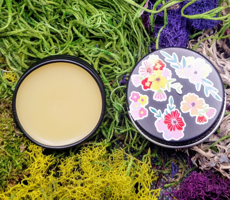 May include: A small round tin with a black lid and a floral design. The lid is open and the tin contains a yellow balm.