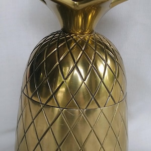 Puede incluir: Un recipiente de metal dorado pulido con forma de piña y tapa extraíble. El cuerpo presenta una superficie texturizada con un patrón de diamantes, y la parte superior está adornada con hojas puntiagudas y estilizadas.