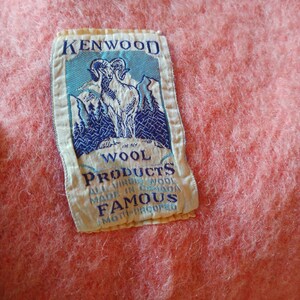 Op de afbeelding: Een roze wollen deken met een wit label waarop staat "Kenwood Wool Products All Virgin Wool Made in Canada Famous Moth-Proofed". Het label toont een blauwe en witte illustratie van een berggeit.