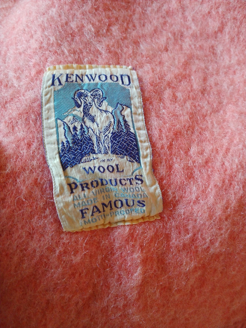 Op de afbeelding: Een roze wollen deken met een wit label waarop staat "Kenwood Wool Products All Virgin Wool Made in Canada Famous Moth-Proofed". Het label toont een blauwe en witte illustratie van een berggeit.