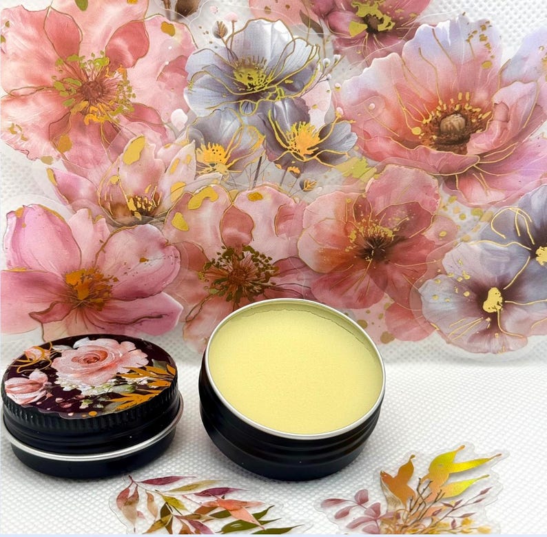 May include: Two small round tins with black lids. The tin on the left has a floral design on the lid. The tin on the right contains a yellow balm. The tins are on a white surface with a floral pattern.