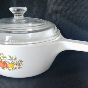 Op de afbeelding: Witte ovenschaal met een glazen deksel en een bloemenmotief met gele, oranje en groene vruchten en bladeren. De schaal heeft een witte handgreep.