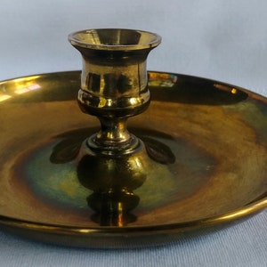 Puede incluir: Un candelero de latón con una base redonda y un solo portavelas. El candelero tiene un asa de anillo en el lateral.