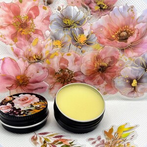 May include: Two small round tins, one with a floral design and the other containing a yellow balm. The tins are on a white surface with a floral background.