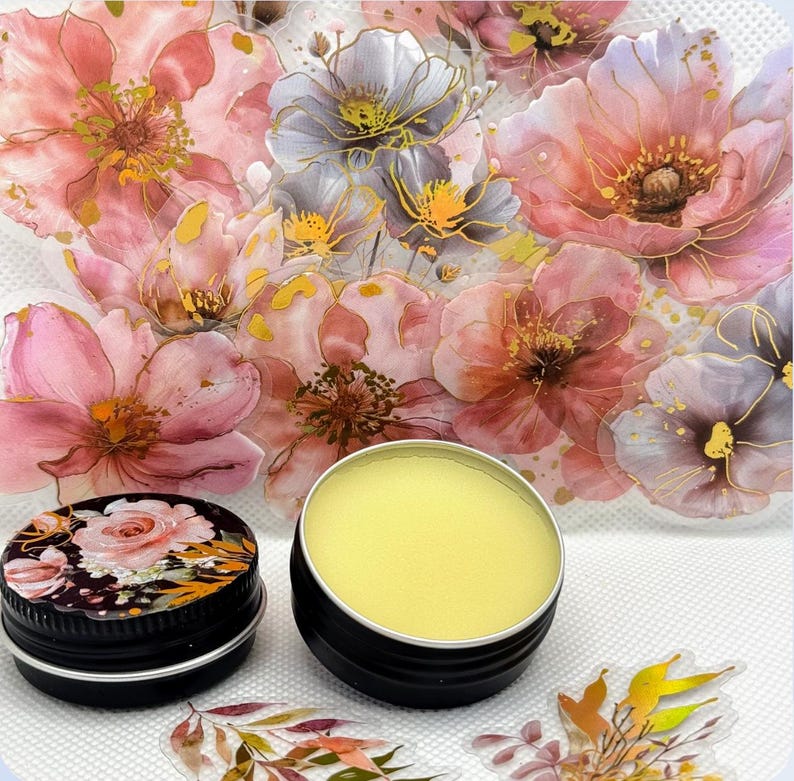 May include: Two small round tins, one with a floral design and the other containing a yellow balm. The tins are on a white surface with a floral background.