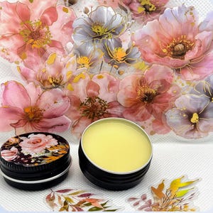 May include: A small, round tin container filled with a yellow balm. The tin is sitting on a white surface with a floral design.