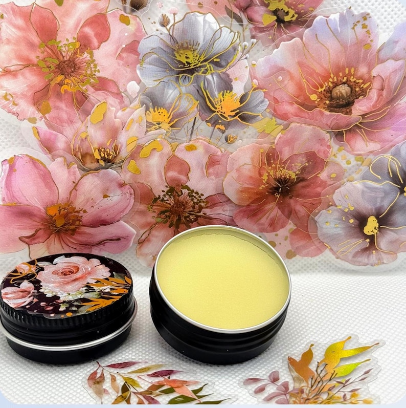 May include: A small, round tin container filled with a yellow balm. The tin is sitting on a white surface with a floral design.