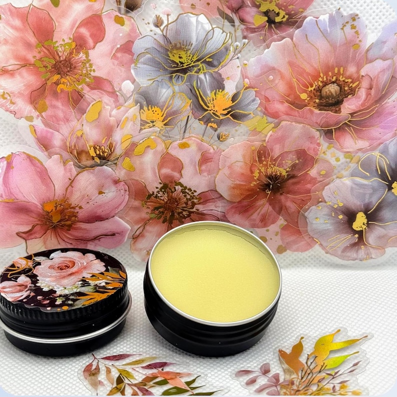 May include: A small tin of yellow lip balm with a floral design on the lid. The tin is sitting on a white surface with a floral background.
