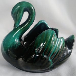 Puede incluir: Un macetero de cerámica en forma de cisne, esmaltado en verde y negro. El cisne tiene un acabado brillante y está en posición enroscada.