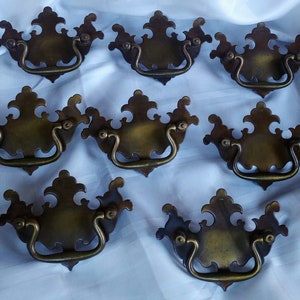 May include: Eight antique brass cabinet pulls with ornate designs. Each pull has a decorative, arched handle.