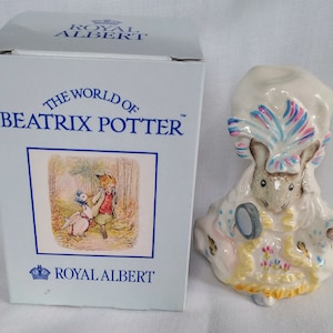 Puede incluir: Una figurita de porcelana blanca de un ratón con un sombrero azul, rosa y blanco y un vestido amarillo. El ratón sostiene un espejo y está sentado sobre una base rosa y amarilla. La figurita forma parte de una colección llamada "El Mundo de Beatrix Potter" de Royal Albert.