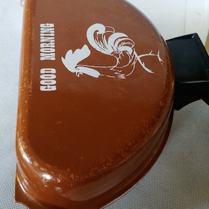 May include: Brown metal container with a white rooster illustration and the words "GOOD MORNING". The container has a rounded shape and a small handle. The rooster design is on the front of the container.