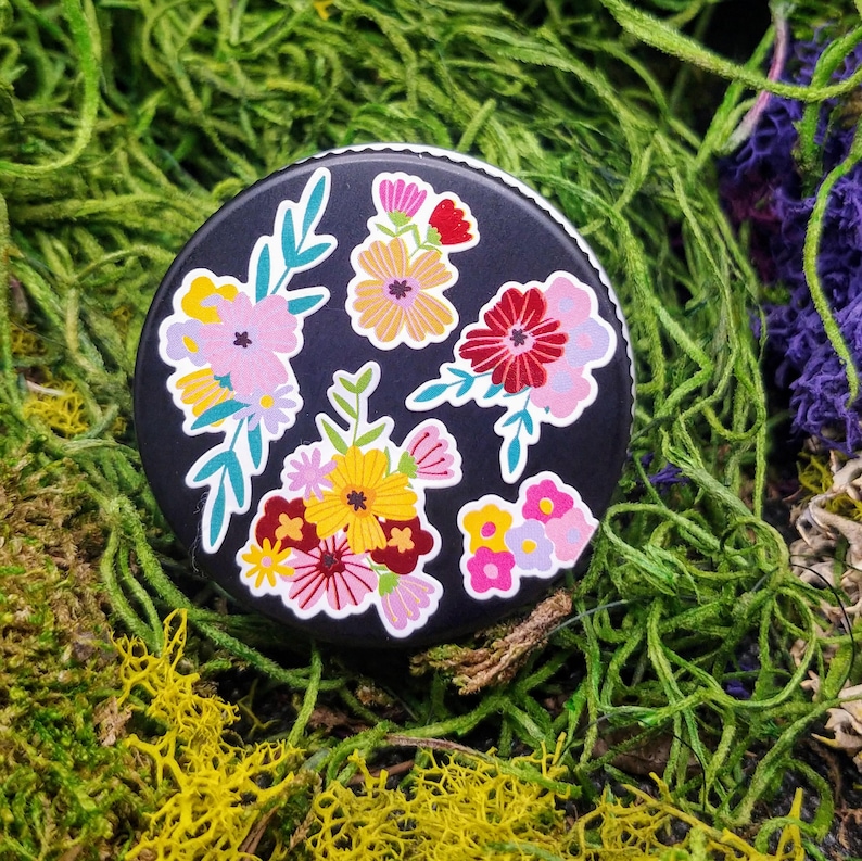 May include: A black round sticker with a floral design. The sticker features pink, red, yellow, and white flowers with green leaves on a black background.