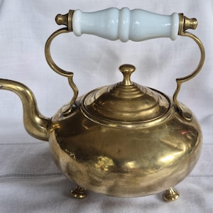May include: A polished brass kettle featuring a rounded body, a curved spout, and a removable lid with a small knob. The kettle stands on three small brass feet and includes a decorative arched handle with a smooth, white ceramic grip.