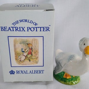 May include: A white porcelain figurine of a goose from the Beatrix Potter collection by Royal Albert. The goose is wearing a blue and white patterned bib and is standing on a green base with yellow flowers.