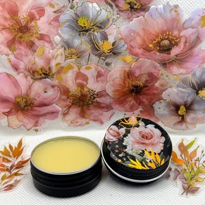 May include: A small tin of yellow lip balm with a floral design on the lid. The lid is black with gold accents and features a pink rose.