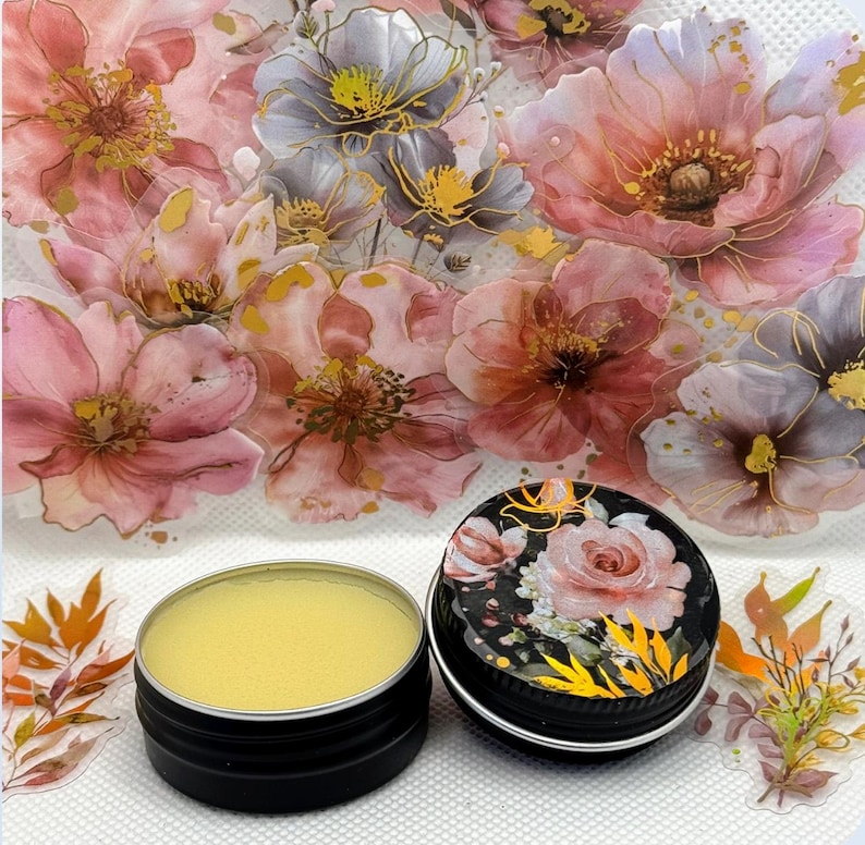 May include: A small tin of yellow lip balm with a floral design on the lid. The lid is black with gold accents and features a pink rose.