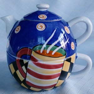 Puede incluir: Una tetera de cerámica azul y blanca con un diseño de taza. La tetera tiene una tapa con un patrón de espiral y la taza tiene rayas rojas, amarillas y blancas.