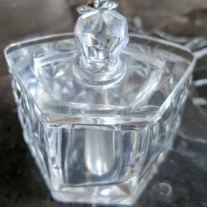 Puede incluir: Un tarro de vidrio transparente con una tapa decorativa que presenta un pomo de cristal. El tarro tiene una forma geométrica y un diseño texturizado.