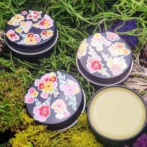 May include: Three small black tins with floral designs on the lids. One tin is open and contains a yellow balm.