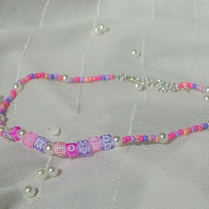 Könnte beinhalten: Ein Perlen-Choker mit den Buchstaben "I AM LOVED" auf quadratischen Buchstabenperlen. Die Halskette hat rosa, lila und weiße Perlen, einen silbernen Verschluss und Perlenakzente. Die Halskette liegt auf einem weißen Hintergrund.
