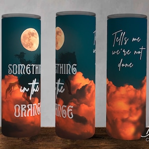 May include: A tumbler with a sunset sky background and a silhouette of a horse and rider. The text "Something in the Orange" is printed on the tumbler. The text "Tells me we're not done" is printed on the other side of the tumbler.