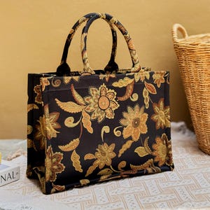 May include: A rectangular tote bag featuring a black base with a repeating golden-yellow and orange floral pattern. The bag has two matching fabric handles, a structured boxy shape, and a flat bottom, showcasing a detailed botanical print design.