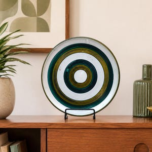 May include: A decorative plate with concentric circles in white, green, and gold tones. The plate is displayed on a black metal stand, showcasing its unique design. The plate is a stylish home decor item.