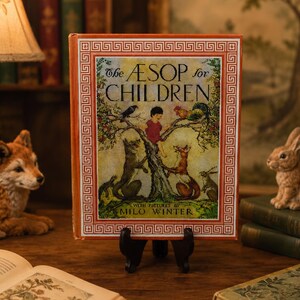 May include: A vintage copy of "The ÆSOP for CHILDREN" book, with a colorful illustration of a child and animals on the cover. The book is displayed on a small stand, with a fox and rabbit figurine nearby. The book has a decorative border.