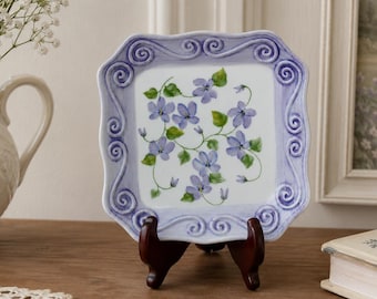 Könnte beinhalten: Ein quadratischer, dekorativer Teller mit lavendelfarbenem Rand und einem Blumendesign aus lila Blumen und grünen Blättern. Der Teller wird auf einem dunkelbraunen Holzständer präsentiert. Der Teller ist ein dekorativer Artikel.
