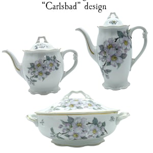 Puede incluir: Una tetera, una jarra de crema y una fuente de servir de porcelana blanca con un diseño floral. El diseño presenta delicadas flores de cornejo rosas y moradas con hojas verdes. El texto "Pirkhammer Dogwood Blossom "Carlsbad" design" está impreso en la parte superior de la imagen.