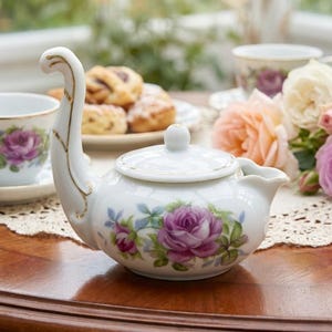 May include: A white porcelain teapot with a curved handle and a lid, decorated with purple rose floral patterns and gold trim. A matching teacup and saucer are visible, along with a plate of pastries and a bouquet of roses.