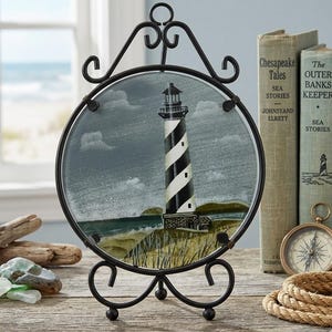 Puede incluir: Una obra de arte de vidrio redondo con un faro de rayas blancas y negras, colocado en un soporte de metal negro. El arte del faro se exhibe sobre una superficie de madera, con libros y una brújula en el fondo.