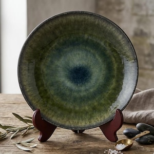 May include: A ceramic plate with a dark green and blue glaze, displayed on a wooden stand. The plate has a circular design with a darker center and lighter edges. The plate is on a wooden table with a sprig of leaves.
