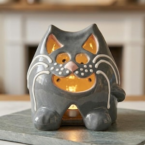 May include: A gray ceramic cat-shaped candle holder with a smiling face, white whiskers, and a pink nose. The candle holder has cut-out eyes and a mouth that glows with the light of a lit candle inside. The cat is sitting on a gray stone surface.