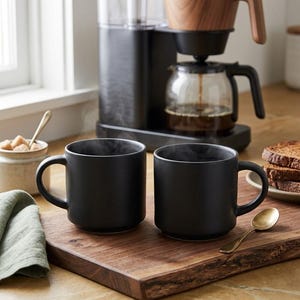 May include: Two black ceramic mugs with steam rising from them sit on a wooden serving board. A coffee maker with a glass carafe and a wooden handle is behind the mugs. A small dish of sugar cubes and a gold spoon are also on the board.
