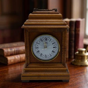 May include: A wooden antique clock with a light-colored face and Roman numerals. The clock is housed in a rectangular wooden frame and sits on a wooden surface. The clock face has a gold-colored trim. The background includes books and a brass inkwell.