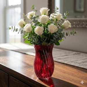 May include: A bouquet of white roses and green eucalyptus leaves in a red glass vase. The vase has a swirled design and sits on a wooden surface. The arrangement is in front of a window.