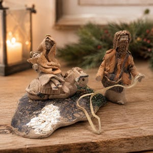 May include: A ceramic nativity scene with three figures and a donkey on a textured base. The figures wear tan and brown robes. The scene is set on a wooden surface with a lit candle and greenery in the background.