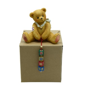 May include: A tan teddy bear figurine with a white collar and a small bell, sitting on a brown cardboard box. The bear holds a string with colorful blocks spelling "NOEL". The bear has red and white accents on its arm.