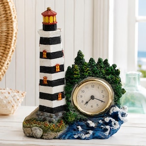 May include: A decorative table clock featuring a tall lighthouse with horizontal black and white stripes, a red roof, and yellow windows. The clock face is circular with a gold rim, set against a base of green trees and blue ocean waves.