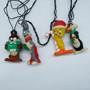 Puede incluir: Una cadena de luces con cuatro figuras de personajes de Looney Tunes: Taz con sombrero de copa y un regalo verde, Bugs Bunny con bufanda roja, Piolín con gorro de Papá Noel y un bastón de caramelo, y Silvestre sosteniendo un regalo.