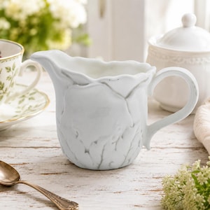 May include: A white ceramic creamer pitcher featuring a sculpted, layered leaf design on its exterior. The pitcher has a curved handle, a wide pouring spout, and a scalloped rim. The surface is smooth and glossy, reflecting light across its textured form.