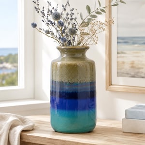 May include: A cylindrical ceramic vase featuring a multi-toned reactive glaze that transitions from a muted olive green at the top to deep navy blue and vibrant turquoise at the base, filled with a dried arrangement of lavender and thistle.