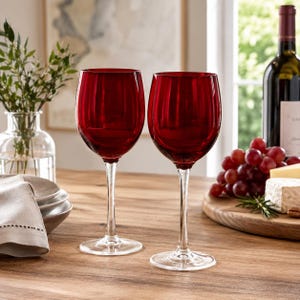 Puede incluir: Dos copas de vino tinto rubí con tallos largos se exhiben sobre una mesa de madera. Una botella de vino, uvas y queso están en el fondo. Las copas están vacías y listas para beber.