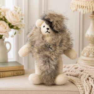Puede incluir: Un oso de peluche crema con una bufanda de piel sintética. El oso lleva un sombrero pequeño y una broche decorativa. La bufanda es una mezcla de gris y marrón. El oso está sobre una superficie beige.