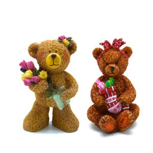 May include: Two decorative teddy bear figurines. One bear holds a bouquet of pink and yellow flowers and a green gift. The other bear wears a red polka dot bow and holds a Christmas stocking with gifts. Both bears have brown fur.
