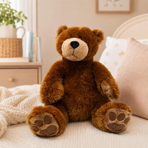 May include: A plush brown teddy bear with a tan muzzle and black nose sits on a bed. The bear has large paws with brown paw pads. The bear is sitting on a white knitted blanket with a pink pillow in the background.