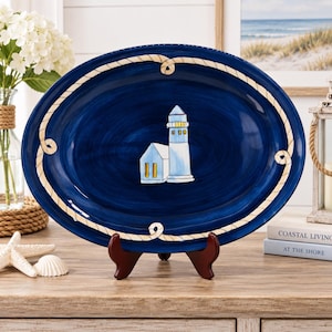 May include: An oval ceramic serving platter with a deep blue finish and a painted white rope border. The center features a lighthouse illustration with a white tower and a small adjacent building, both accented with yellow windows and blue shading.