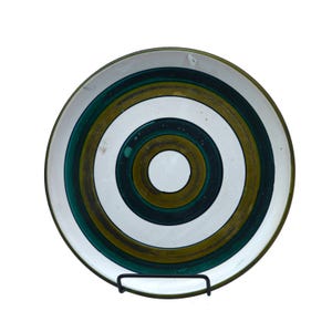 May include: A circular ceramic plate featuring a design of concentric rings in white, green, and gold. The plate has a dark green rim and is displayed on a black metal stand. The design is centered, creating a bold, graphic look.